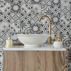 May include: A bathroom vanity featuring a white ceramic sink and a gold tap. The vanity has a wooden cabinet and a white countertop. The background showcases patterned tiles in grey and white. A candle and a small container are on the countertop.