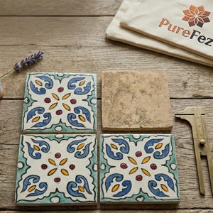 May include: Four decorative square tiles with a floral design in blue, yellow, and red on a white background. One plain tile, a brass caliper, a brush, and a canvas bag with the text "PureFez" are also present.