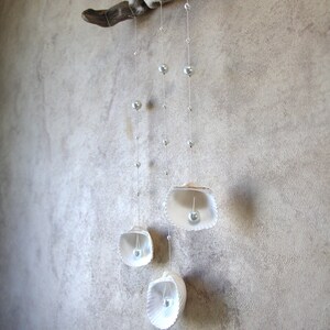 Puede incluir: Un móvil de madera flotante blanco con tres conchas blancas colgando de él. Cada concha tiene una perla en su interior. El móvil está colgado de una pared blanca.