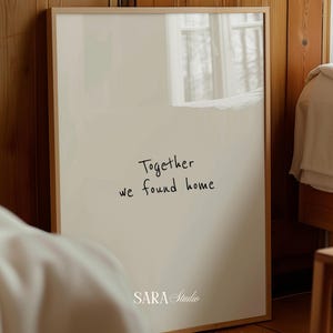 Può includere: Una stampa incorniciata con sfondo beige chiaro e il testo scritto a mano "Together we found home". La stampa è in una cornice di legno chiaro e presenta il logo "SARA Studio" in basso. La stampa è appoggiata a una parete di legno.