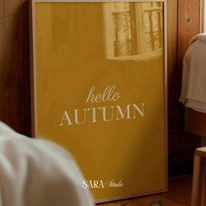 May include: A rectangular wall art print in a light wood frame featuring a solid mustard yellow background with the white text hello AUTUMN centred and the white text SARA Studio printed at the bottom centre of the poster.