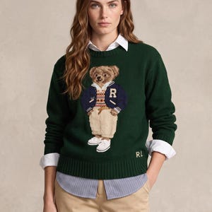 May include: A dark green long-sleeve knit sweater featuring a central graphic of a teddy bear wearing a navy blue varsity jacket, a patterned sweater vest, and tan trousers. The letters RL are embroidered in gold on the lower right side.