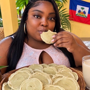 May include: A light pink sleeveless top with a keyhole neckline and sequin detailing, paired with a wooden bowl filled with numerous round, pale green cookies, and a glass of beige liquid, with a Haitian flag visible in the background.