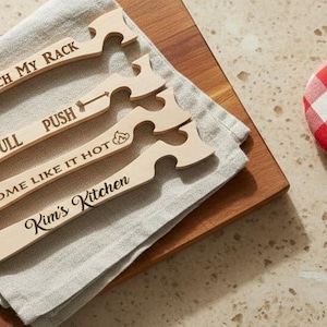 May include: A kitchen scene with wooden serving utensils, a mug of coffee, and chocolate chip cookies on a cooling rack. A red and white gingham oven mitt is also present. The utensils have text on them.