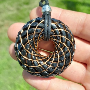 Peut inclure: Un pendentif circulaire composé d'un noyau noir texturé en forme de beignet, enveloppé de fins fils métalliques dorés disposés en spirale. Le pendentif est suspendu à un cordon noir fixé par une petite perle en métal argenté au sommet.