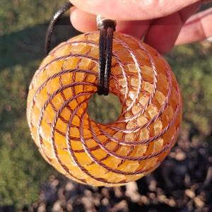Pode incluir: Um colar com pingente em forma de donut. O pingente é feito de pedras laranja facetadas, envoltas em fio de cobre. O colar tem um cordão preto. O pingente é segurado contra um fundo verde desfocado.