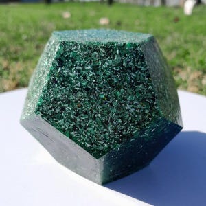 Puede incluir: Geoda de cristal verde oscuro facetada, con forma geométrica. La superficie es un mosaico de pequeños fragmentos verdes brillantes, creando una apariencia texturizada. El cristal está sobre una superficie blanca con un fondo verde borroso.