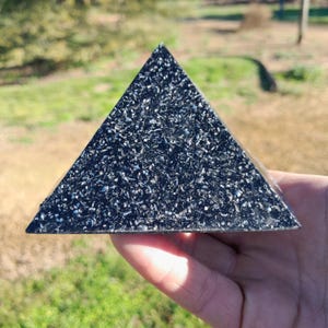 以下が含まれることがあります： キラキラとした粒状の表面を持つ、暗く質感のあるピラミッド。 幾何学的な形状は手で持たれており、背景はぼやけた緑と茶色です。