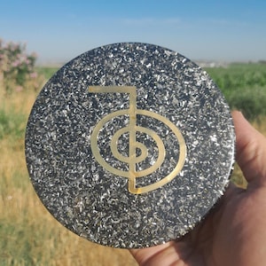 May include: A round, dark gray orgonite disc with a gold-colored Reiki symbol in the center. The disc has a textured surface and is held in a person's hand. The background shows a field and a blue sky.