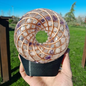 May include: A clear sphere filled with white and purple stones, encased in copper wire, and mounted on a black base. The sphere has a spiral design with a central opening. The background is a sunny outdoor scene.