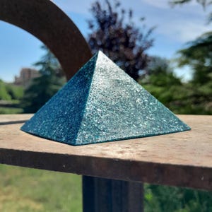 May include: A teal pyramid object with a textured, glittery surface. The pyramid has four triangular faces meeting at a sharp apex, featuring a dense, sparkling finish that reflects light across its geometric, multi-faceted, and solid structure.