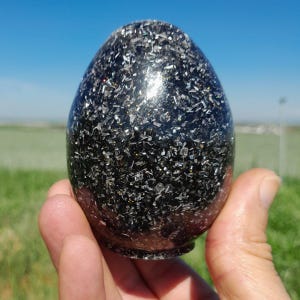 Puede incluir: Un objeto decorativo negro en forma de huevo con una superficie texturizada y brillante. El objeto se sostiene en una mano sobre un fondo de cielo azul y campo verde. La superficie está cubierta de pequeños trozos reflectantes.