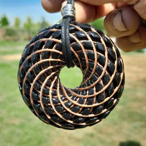 May include: A circular pendant featuring a woven pattern of glossy black segments intertwined with thin copper-colored wire. The pendant is suspended from a black cord that passes through a silver-toned metal bead at the top of the circular frame.
