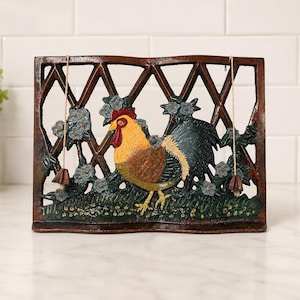 May include: A rectangular cast iron trivet featuring a central cockerel with a yellow head, red comb, and dark tail feathers. The cockerel stands on a green grassy base against a brown lattice fence backdrop with blue flowers and two hanging charms.