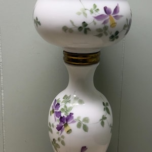 May include: A white glass oil lamp featuring hand-painted purple flowers and green leaves on the base and font. The lamp has a brass burner assembly with a black knob and a dark, ornate, cast metal base with intricate cut-out leaf patterns.