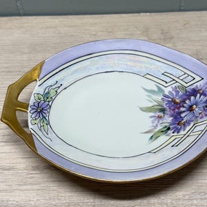 May include: A leaf-shaped porcelain serving dish with a light purple border and a central white area. The dish features a painted cluster of purple daisies with green leaves, gold-painted handle accents, and gold trim along the outer edge.