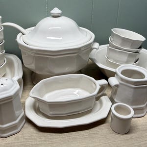 Puede incluir: Un juego de vajilla de cerámica blanca que incluye cuencos, tazas, una sopera con tapa, una salsera, saleros y pimenteros, y jarras. El conjunto tiene un diseño clásico, ideal para servir comidas.