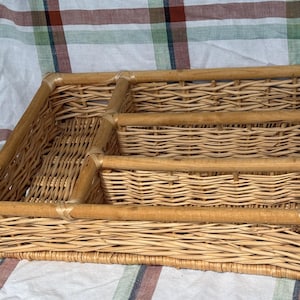 May include: A rectangular light brown wicker tray featuring a sturdy bamboo frame and three distinct interior compartments. The woven basket is constructed with natural rattan reeds, showcasing a detailed interlaced pattern throughout the base and sides.
