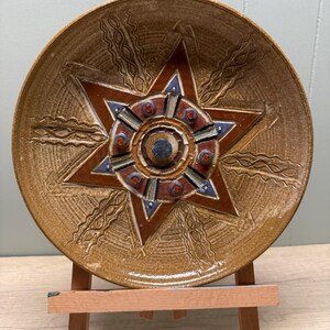 Può includere: Un piatto decorativo in ceramica con un motivo a stella. Il piatto presenta una base marrone con una stella in rilievo al centro, accentuata da dettagli rossi, blu e arancioni. Il piatto è esposto su un piccolo cavalletto di legno.