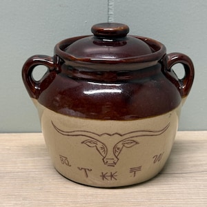 Puede incluir: Un tarro de cerámica marrón y beige con tapa y dos asas. El tarro presenta un diseño de cuerno largo y letras grabadas. La tapa y la parte superior son de color marrón brillante.