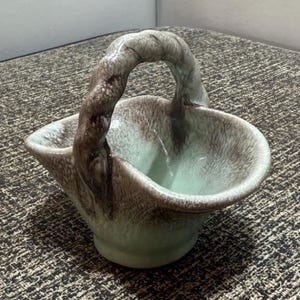 Pode incluir: Uma pequena cesta de cerâmica com uma alça. A cesta é verde claro com detalhes em marrom e uma alça texturizada. A cesta curva é provavelmente para uso decorativo. Medidas em centímetros.