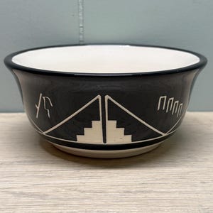 May include: A black ceramic bowl with a white interior and a black rim. The exterior features a geometric design in white, including triangles and stepped patterns. The bowl sits on a light-coloured wooden surface.
