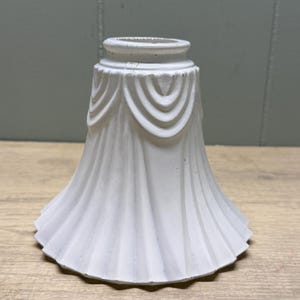 May include: A white lamp shade with a flared, bell-like shape and a matte finish. The exterior features a series of vertical pleats or ridges that extend from the base to the top, where they are accented by a decorative draped swag pattern.