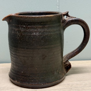 Puede incluir: Jarra de cerámica marrón oscuro hecha a mano, con pico vertedor y asa grande. La jarra tiene un aspecto rústico y texturizado, con variaciones de color y un elemento de diseño único cerca del asa.