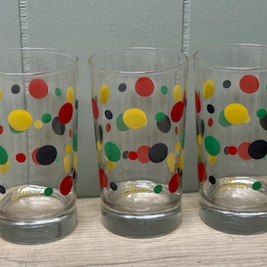 Puede incluir: Tres vasos transparentes con base gris, decorados con lunares de colores rojo, amarillo, verde y negro. La palabra "Fiesta" está impresa en la parte delantera de cada vaso. Los vasos son cilíndricos y parecen ser de vidrio.