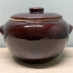 May include: A brown ceramic casserole dish with a lid. The dish has two handles and a small knob on the lid. The exterior has a glossy finish. The dish is sitting on a light-colored wooden surface.