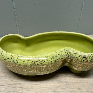 May include: A vintage, oblong planter with a speckled, lime green exterior and a smooth, lighter green interior. The planter has a unique, double-lobed design and gold-coloured accents, ideal for small plants or succulents.