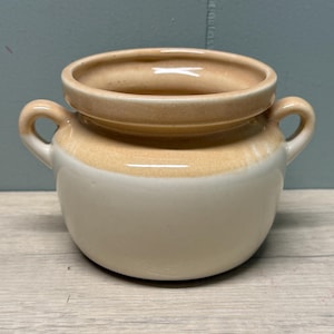 May include: A small, round ceramic pot featuring two side handles. The upper portion and rim are glazed in a warm, light tan color, while the lower body is finished in a smooth, creamy off-white glaze. The vessel has a wide, open mouth.