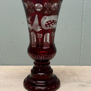 May include: A ruby red glass vase with etched white designs. The vase features a flared top, a bulbous midsection, and a pedestal base. The etched designs include swirls, circles, and stylized animal figures. The vase is displayed on a wooden surface.