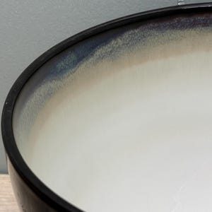 May include: Close-up of a ceramic bowl with a black rim. The bowl's interior features a gradient of colors, transitioning from white to beige and then to blue near the rim. The bowl is likely for serving or display.