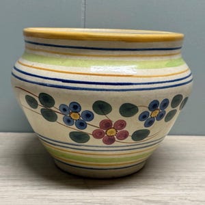 May include: A cream-coloured ceramic pot with yellow, green, and blue horizontal stripes. It features hand-painted floral designs in blue and red, with green leaves. The rim is yellow. A decorative item.