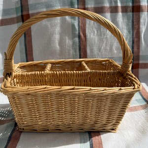 May include: A rectangular light brown woven wicker basket featuring a single arched handle and an interior divided into three separate compartments. The basket is constructed from natural reed material with a tight, uniform weave pattern throughout.