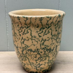 Puede incluir: Un jarrón de cerámica beige con un patrón moteado verde. El jarrón tiene un borde ligeramente ensanchado y un cuerpo redondeado. El diseño moteado cubre toda la superficie, creando una apariencia única y rústica. Adecuado para flores o como pieza decorativa. Aproximadamente 15 cm de alto.