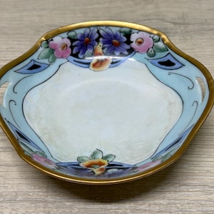 May include: A small, shallow porcelain dish with a scalloped edge and gold trim. The interior features a light blue border decorated with hand-painted purple and pink flowers, green leaves, and black geometric accents against a white center.