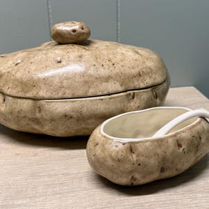 May include: A ceramic potato-shaped serving dish with a lid and a small matching serving bowl with a spoon. The set is beige with brown speckles, resembling a real potato. The lid has a small potato-shaped knob.