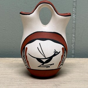 May include: A ceramic vessel featuring a bridge handle and two openings. The exterior is painted with a cream base, accented by terracotta red bands and black line work. A stylized black bird graphic is centered on the front of the rounded body.
