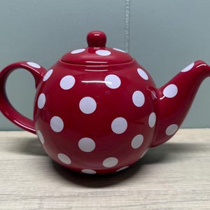 May include: A red ceramic teapot with white polka dots. The teapot has a curved handle, a spout, and a lid with a small knob. The teapot is sitting on a light-colored wooden surface.