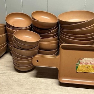 Pode incluir: Uma coleção de tigelas de servir marrons e uma bandeja retangular com uma alça. As tigelas são empilhadas em várias pilhas, de tamanhos variados. A bandeja tem um rótulo que diz "Barbecue Tray".