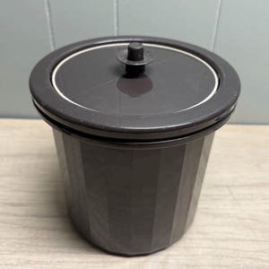 Può includere: Un contenitore marrone scuro con coperchio, di forma cilindrica e con esterno scanalato. Il coperchio ha una piccola maniglia e un anello bianco. Il contenitore è probabilmente in plastica ed è adatto allo stoccaggio.