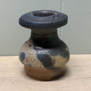 May include: A small ceramic vase with a wide, flat rim and a bulbous body. The piece features a mottled glaze with dark navy blue, black, and speckled tan tones. The base is unglazed, revealing the natural brown clay texture of the pottery.
