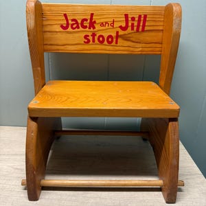Op de afbeelding: Een houten kruk met een natuurlijke lichtbruine afwerking en een rugleuning. Op de rugleuning staat de rood geschilderde tekst "Jack and Jill stool". De kruk heeft een platte zitting, twee zijsteunen en een horizontale houten verbindingsstang.