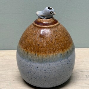 May include: A ceramic vessel with a rounded, egg-like shape featuring a smooth, glossy glaze. The bottom half is a speckled light blue, transitioning into a rich, warm brown at the top. A small, light blue sculpted bird sits atop the vessel.
