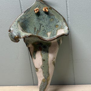 May include: A cone-shaped ceramic wall planter featuring a mottled green and white glaze with a glossy finish. The vessel has a wide, flared opening with an irregular rim and includes two small holes at the top threaded with a tan leather cord.