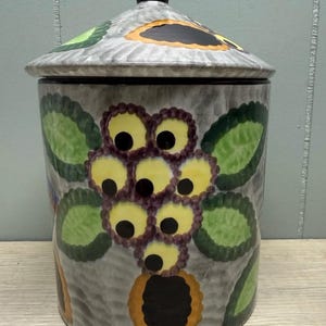 Może przedstawiać: Ceramiczny słoik z pokrywką, szara podstawa z teksturowanym wzorem. Ozdobiony kolorowymi wzorami kwiatowymi w kolorze żółtym, zielonym i pomarańczowym. Pokrywka ma czarny uchwyt i pasujące akcenty kwiatowe.