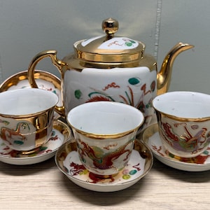 Puede incluir: Un juego de té de porcelana blanca con ribete dorado, que incluye una tetera, tazas y platillos. El juego está decorado con coloridos diseños de dragones y flores. La tetera tiene un asa y una tapa doradas.