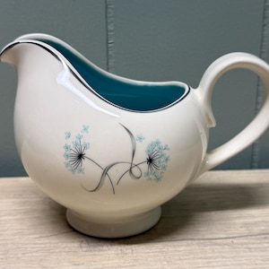 Peut inclure: Un pot à lait en céramique de couleur crème avec une anse incurvée et un bec verseur. L'intérieur est d'un turquoise vif. L'extérieur présente un motif délicat de deux fleurs stylisées ressemblant à des pissenlits en bleu clair et noir.